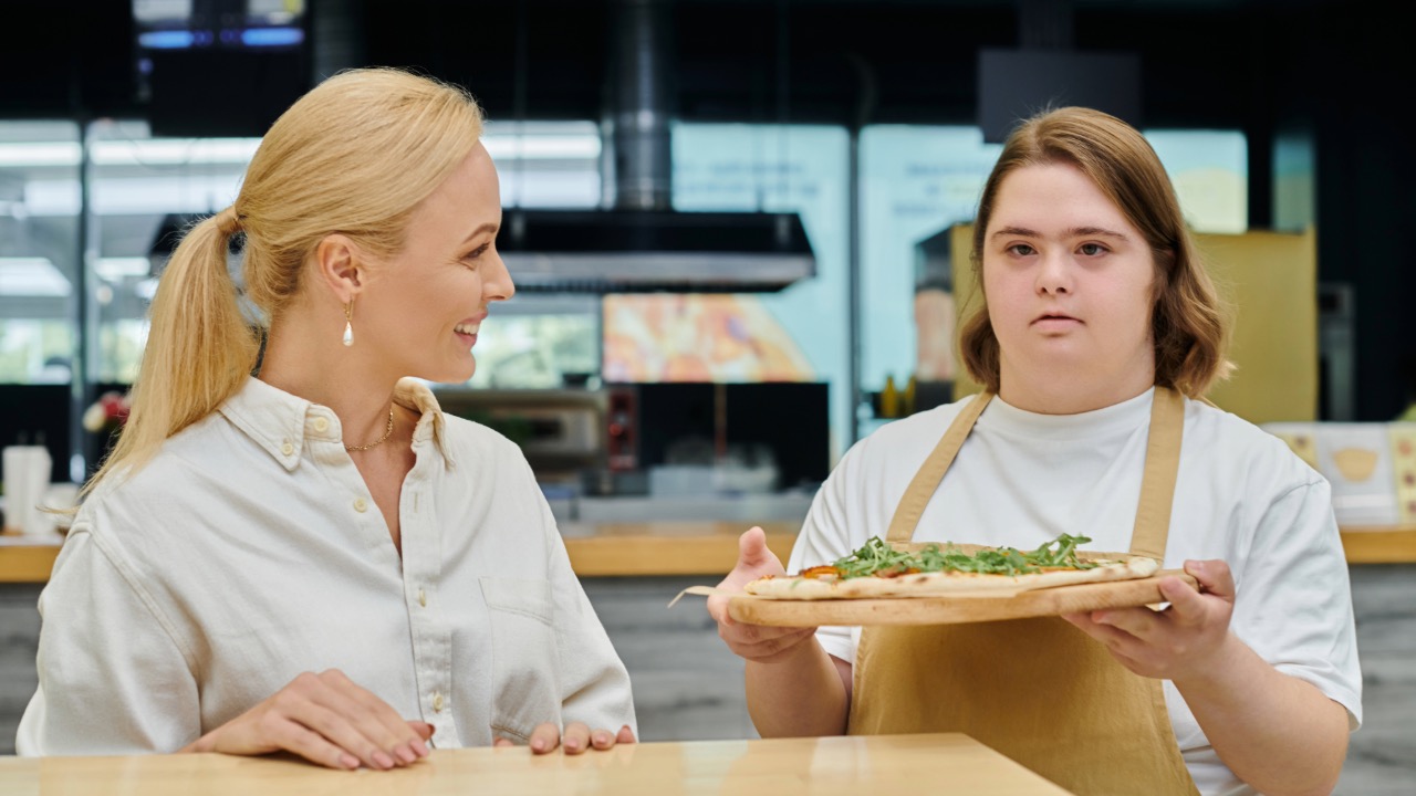 two employees at a pizza restaurant one holding pizza has special needs